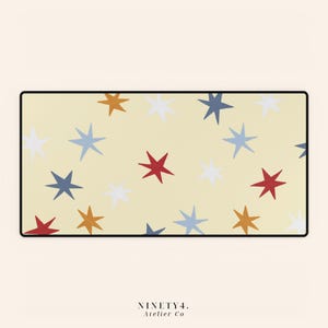 May include: A rectangular desk mat with a pale yellow background featuring a scattered pattern of six-pointed stars in shades of red, white, blue, and mustard yellow. The mat has a thin black border and the text NINETY4. Atelier Co at the bottom.