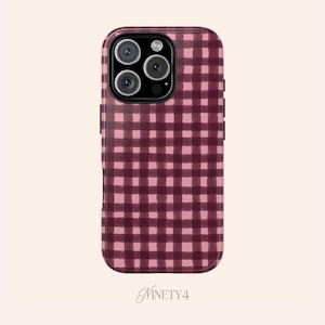 Peut inclure: Coque de téléphone avec un motif vichy bordeaux et rose. L'étui a une finition brillante et un module caméra avec trois objectifs. La marque "NINETY4" est en bas.