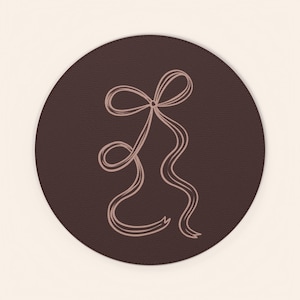 May include: A brown, circular coaster with a light pink line drawing of a bow. The bow has two loops and long, flowing ribbons. The coaster is a solid, dark brown color.