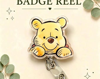 May include: A badge reel featuring a yellow bear character with a brown nose and smiling expression. The bear is rendered in a watercolour style with a glittery acrylic finish. The text BADGE REEL is printed above the character in dark green.