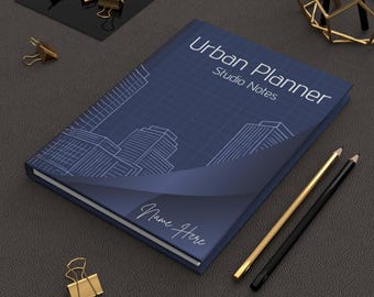 Może przedstawiać: Granatowy notatnik Urban Planner Studio Notes z projektem miasta w stylu planu. Zawiera złote i czarne ołówki, złote spinacze do papieru i geometryczną złotą rzeźbę z drutu na ciemnoszarej powierzchni.