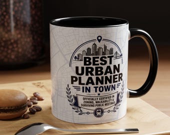 Może przedstawiać: Biały ceramiczny kubek z czarnym wnętrzem i uchwytem. Kubek zawiera napis "BEST URBAN PLANNER IN TOWN" w granatowym wzorze z tłem krajobrazu miejskiego i mapy. Zawiera tekst "OFFICIALLY CERTIFIED IN ZONING, WALKABILITY & SURVIVING PUBLIC MEETINGS."