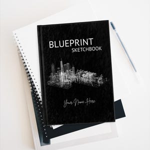 Op de afbeelding: Een zwart Blueprint schetsboek met een witte skyline. De omslag heeft de tekst "Blueprint Sketchbook" en "Your Name Here" in handschrift. Het ligt op een wit spiraalgebonden notitieboek.