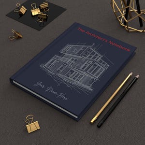 Op de afbeelding: Een marineblauw notitieboek met de titel "The Architect's Notebook" met een gedetailleerde architectonische schets op de omslag. Het notitieboek bevat de tekst "Your Name Here" en wordt vergezeld door gouden en zwarte potloden en gouden paperclips.