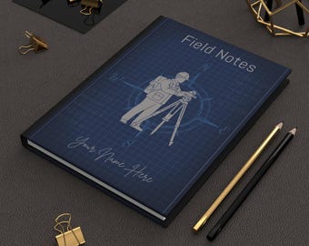 Może przedstawiać: Niebieski notatnik z napisem "Field Notes" i "Your Name Here" na okładce. Okładka ma wzór planu z grafiką geodety i różą wiatrów. Widoczne są również złote i czarne ołówki oraz złote spinacze do papieru.