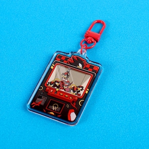 May include: A red and black acrylic keychain featuring a cartoon image of Shadow the Hedgehog from the Sonic the Hedgehog video game series. The keychain is shaped like a claw machine with Shadow and several smaller versions of himself inside. The keychain has a red metal clasp.