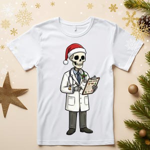 Puede incluir: Camiseta blanca con un dibujo de un esqueleto de médico con gorro de Papá Noel, bata de laboratorio y estetoscopio. El esqueleto sostiene una lista con el texto "NOEL LISTES". Diseño humorístico navideño.
