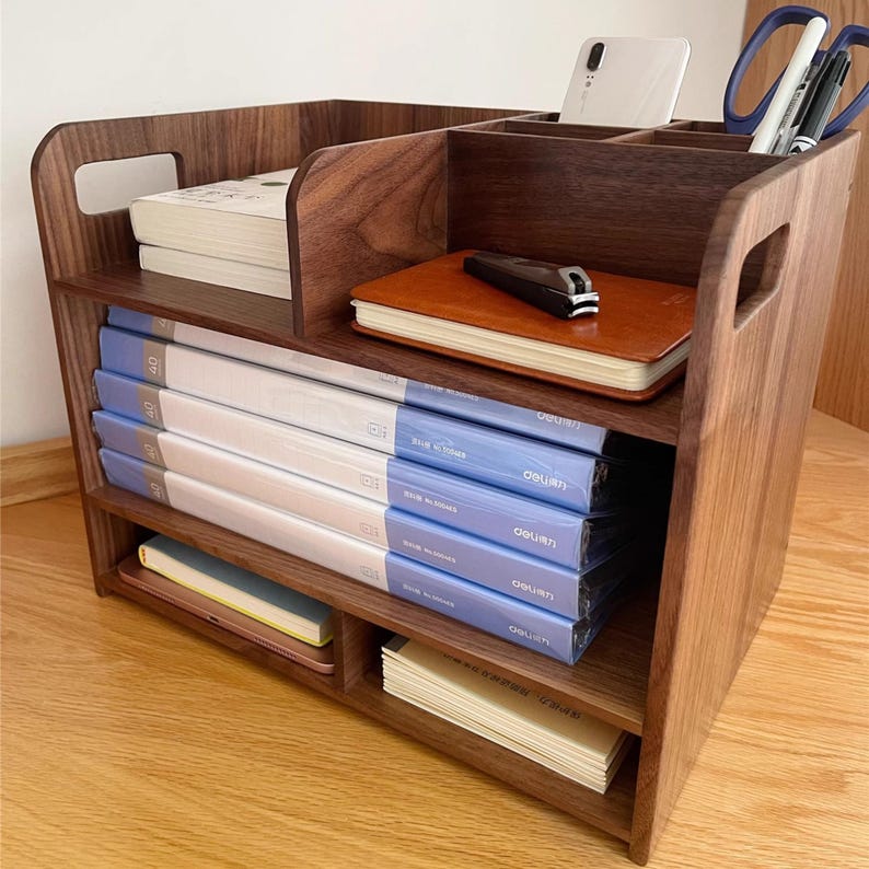 Op de afbeelding: Een houten bureau-organizer met meerdere compartimenten. Het bevat boeken, notitieboekjes en mappen met blauwe en witte covers. Een smartphone, schaar en pennen zijn ook zichtbaar. De organizer is van donkerbruin hout.