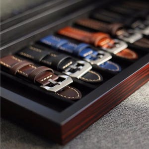May include: A set of six leather watch straps in a wooden box. The straps are in various colours, including brown, black, blue, and orange. Each strap has a metal buckle.