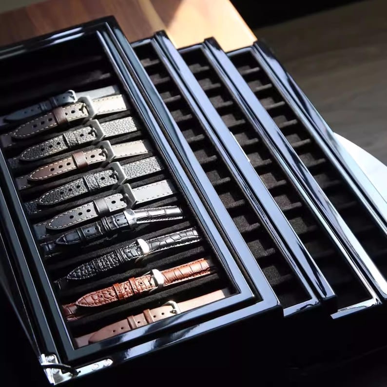 May include: A black wooden watch box with multiple layers and compartments for storing watch straps. The box is open, revealing a variety of watch straps in different colours and materials.