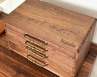 May include: A wooden jewelry box with three drawers and brass-colored handles. The box is a warm brown color with visible wood grain. The name "Natalie" is engraved on the top right corner of the box.
