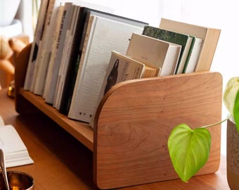 May include: A wooden book organizer with a curved front and two shelves. The organizer is filled with books and is sitting on a wooden desk. A green plant is in the background.