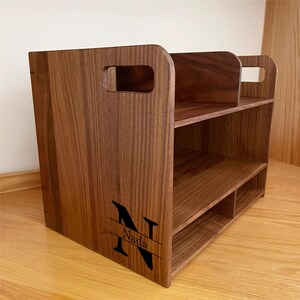 Op de afbeelding: Een houten bureau-organizer met een natuurlijke houtnerf afwerking. De organizer heeft twee planken en een verdeeld onderste gedeelte. Het heeft aan elke kant een handvat en de naam "Nada" met de letter "N" gegraveerd op de voorkant.
