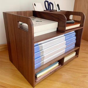 Op de afbeelding: Een donkerbruine houten bureau-organizer met meerdere compartimenten en planken. Het bevat boeken, notitieboekjes, dossiers, een smartphone, pennen en een schaar. De organizer heeft een handvat aan de zijkant.