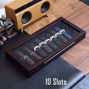 May include: A dark brown wooden watch strap storage box with a clear glass top. The box has 10 slots for storing watch straps and the text "10 Slots" is printed on the bottom of the box.