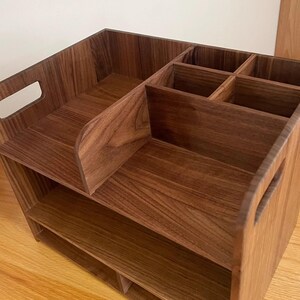 Op de afbeelding: Een houten bureau-organizer in donkerbruin met meerdere compartimenten en planken. De houtnerf is zichtbaar. Het heeft een handvat aan de zijkant voor gemakkelijk dragen. Ontworpen voor het opbergen van kantoorbenodigdheden of andere items.