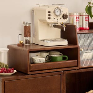 Puede incluir: Una máquina de espresso color crema con un manómetro y un mango de madera se encuentra sobre un mueble de madera oscura. Un molinillo de pimienta de madera y latas de café están cerca. El mueble tiene un estante con tazas y un cuenco con el texto "ANN HUNTING".