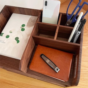 Op de afbeelding: Een houten bureau-organizer met meerdere compartimenten. Het bevat een smartphone, notitieboek, pennen, schaar en een klein bruin lederen notitieboek met een nagelknipper. De organizer is gemaakt van donker hout.
