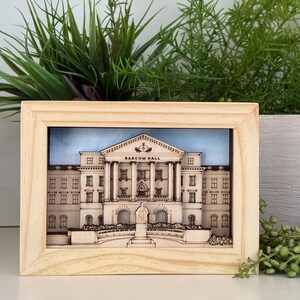May include: A wooden framed picture of a building with the text "Bascom Hall" above the entrance. The building is a light brown color with white trim. The picture is set against a blue sky background.