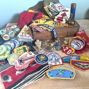 May include: A collection of vintage Boy Scout and military memorabilia, including patches, pins, and a flashlight. The patches feature text like "U.S. Air Force," "Suryan Man," and "Netseo Trails Council BSA." The items are arranged on a wooden surface.
