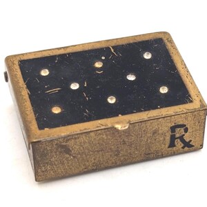May include: A small, rectangular antique pillbox with a black top and gold-colored sides. The top features eight white and gold dots. The letter "R" is printed on the side of the box.