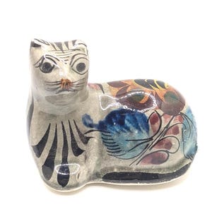 May include: A ceramic cat figurine with a white base and black and blue painted details. The cat is lying down with its head turned to the side. The figurine is decorated with colorful floral patterns.
