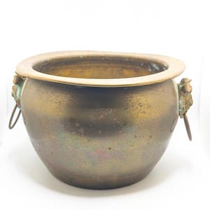 May include: A round, antique brass pot with a wide rim and two decorative lion's head handles. The pot has a weathered, aged appearance with a dark, mottled finish. The pot is likely used for decorative purposes.
