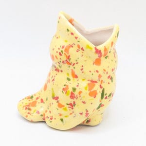 May include: A small, yellow ceramic boot-shaped vase with a speckled pattern of orange, red, and green. The vase has a wide opening and a smooth, glossy finish. The boot is a decorative item.