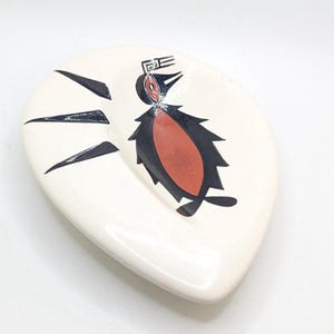 May include: White ceramic decorative dish with a stylized penguin design. The penguin is depicted in black and red, with black arrow-like shapes. The dish has a smooth, glossy finish and a unique, organic shape.