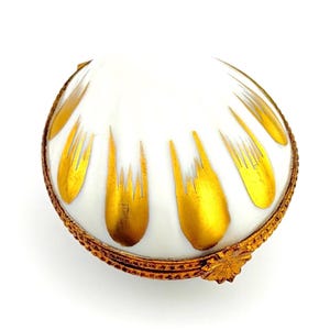 May include: A white, shell-shaped trinket box with gold accents. The box features a gold-colored rim and a decorative gold clasp. The top of the box has a pattern of gold leaf-like designs radiating from the center.