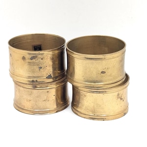 May include: Four vintage brass napkin rings. Each ring is cylindrical with a slightly aged, tarnished appearance. The rings are stacked in pairs, showcasing their simple, classic design. Perfect for adding a touch of elegance to any table setting.