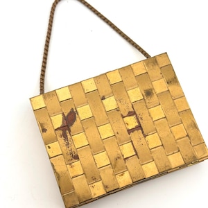 May include: A vintage gold-toned compact with a woven pattern, featuring a small brown chain handle. The compact has some discoloration, adding to its antique charm. The square shape is classic and elegant.