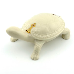 May include: A cream-colored ceramic turtle figurine. The turtle has a textured shell with a decorative gold and red accent. The figurine is on four short legs and has a long neck and head.