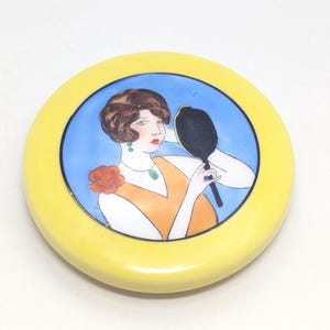 May include: A round, yellow compact with a painted illustration of a woman looking in a hand mirror. The woman is wearing an orange dress, a green necklace, and a red rose. The background is blue.