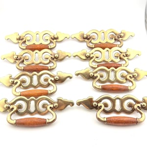 May include: Eight vintage brass drawer pulls with wooden handles. The brass hardware has an ornate, scrolled design. The wooden handles are a warm brown color. These pulls are likely for a dresser or cabinet.