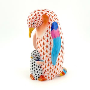 May include: A ceramic penguin figurine with a parent and baby penguin. The parent penguin has a white and orange patterned body, a yellow beak, and blue and pink wing accents. The baby penguin is black and white.