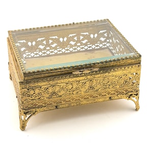May include: A rectangular, gold-toned jewelry box with a clear glass top. The box features ornate floral and scrollwork detailing on the sides and legs. The hinged lid has a decorative filigree design. The box is elevated on four curved feet.
