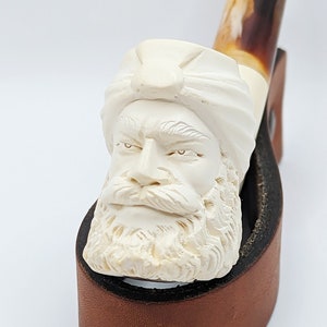 May include: A white ceramic pipe with a carved face wearing a turban. The pipe is resting on a brown leather stand.