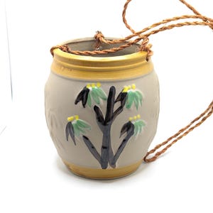 May include: A ceramic hanging planter with a beige background and a brown, black, and green floral design. The planter has a yellow rim and a rope hanger.