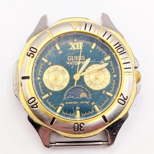 Op de afbeelding: Een Guess-horloge in twee tinten, goud en zilver, met een blauwe wijzerplaat. Het horloge heeft een dag- en datumaanduiding, evenals een maanfase-indicator. Het horloge is waterdicht tot 50 meter of 165 voet.