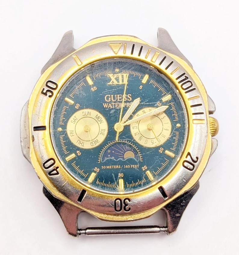 Op de afbeelding: Een Guess-horloge in twee tinten, goud en zilver, met een blauwe wijzerplaat. Het horloge heeft een dag- en datumaanduiding, evenals een maanfase-indicator. Het horloge is waterdicht tot 50 meter of 165 voet.