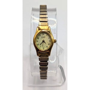 May include: A gold-toned Sharp Quartz wristwatch with a cream-colored face and black numerals. The watch has a stretch band and a small, oval-shaped face. The watch is displayed on a clear, curved stand.