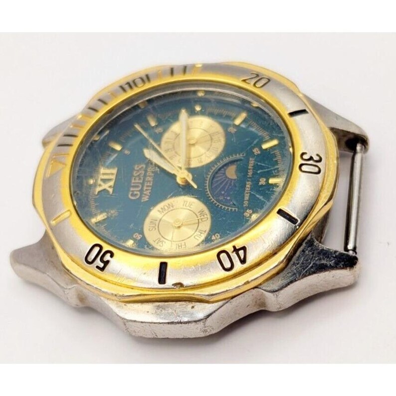 Op de afbeelding: Een gouden en zilveren Guess-horloge met een blauwe wijzerplaat en een datumavenster. Het horloge heeft een maanfase-complicatie en een dag van de week-indicator.