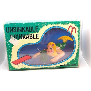 May include: A box for an "Unsinkable Drinkable" mermaid toy. The mermaid is depicted in a blue pool with a diving board and a red cup. The box is green with white text and a yellow background for the pool.
