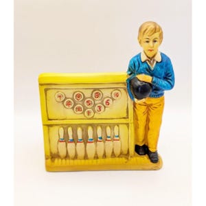 May include: A ceramic figurine of a boy holding a bowling ball, standing next to a yellow bowling alley with ten pins. The pins are numbered 1 through 10.