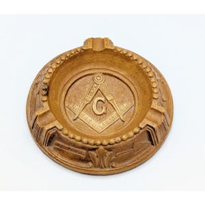 May include: A round, wooden ashtray with a carved Masonic symbol in the center. The symbol features a compass, square, and the letter "G". The ashtray has a light brown color and a decorative border.