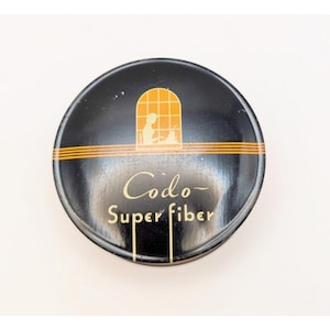 May include: A round, black tin with the words "Codo Super Fiber" in gold lettering. The tin has a gold stripe and a gold and orange illustration of a person working. The tin is likely vintage and used for a hair product.