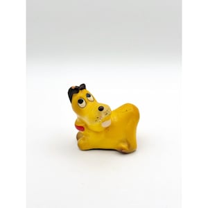 May include: A yellow dog figurine with black ears, a red collar, and large white eyes with black pupils. The dog is in a seated position, looking upward with a light-colored muzzle and small black dots representing whiskers on its snout.