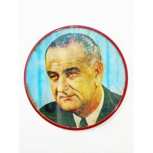 May include: A round pin featuring a portrait of a man in a suit and tie. The portrait is set against a blue background with vertical lines and is framed by a red border. The image is a close-up.
