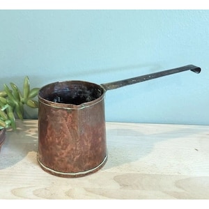 May include: A copper-colored, antique-style pot with a long, slender handle. The pot has a cylindrical body with a slightly flared rim and a white metal band around the base. The handle is made of a darker metal and curves slightly at the end.